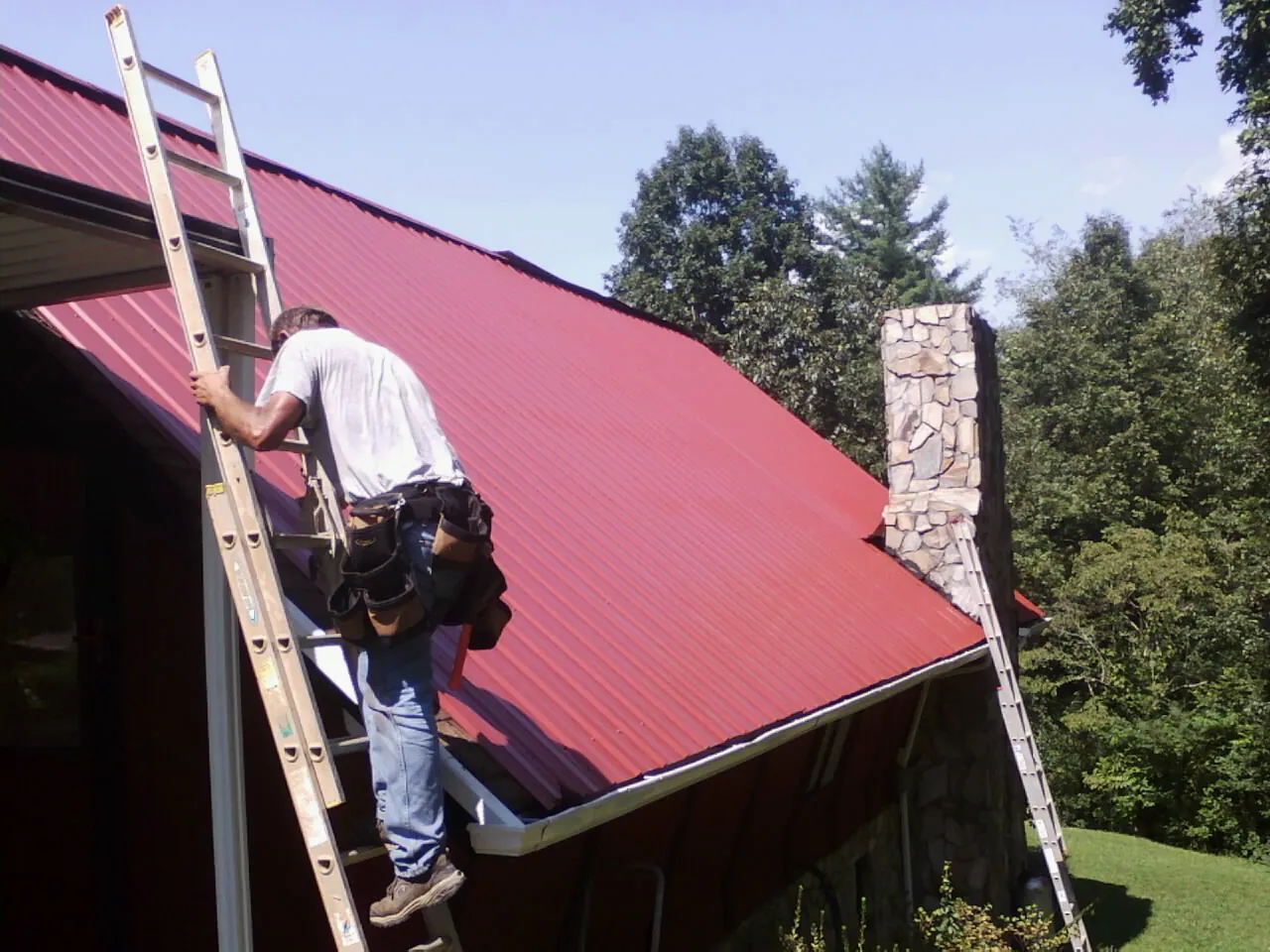 Roofer on a ladder beside a red metal roof and stone chimney — Soffit & Fascia Repair by Roofing Brothers in Stroudsburg